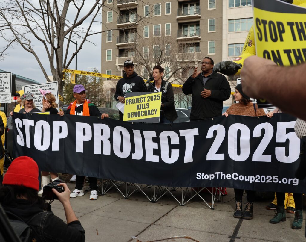Menschen bei einem Protest hinter einem Banner mit der Aufschrift: Stop Project 2025. Schild mit Aufschrift: "Project 2025 Kills Democracy"