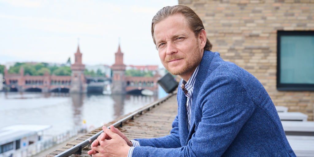 Sebastian Klein lehnt an eine Mauer, im Hintergrund sieht man verschwommen einen Fluss und eine Stadt